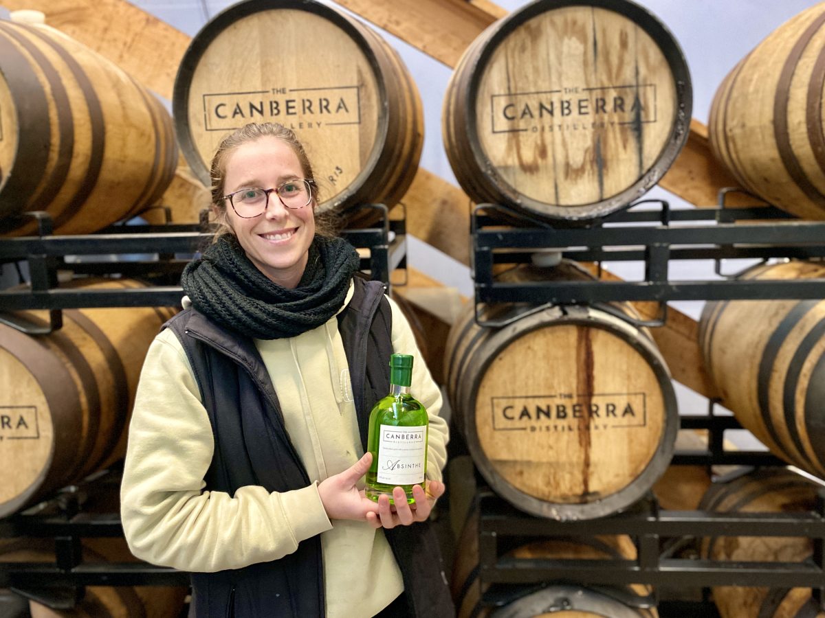 woman holds a bottle of green spirit with Canberra Distillery branded barrels behind her.