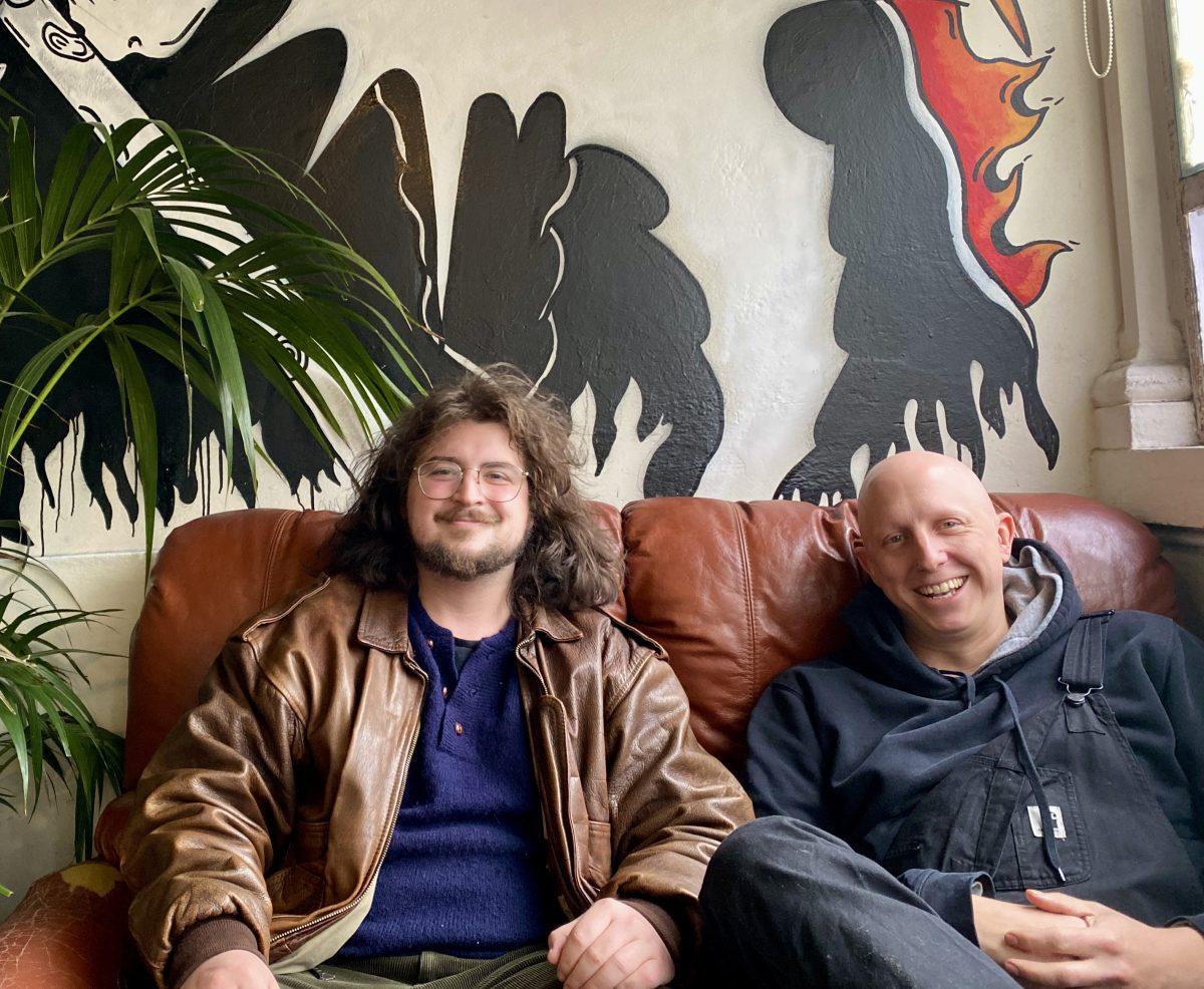 Two men sit side by side on a brown leather couch next to an indoor plant. There is a mural on the wall.