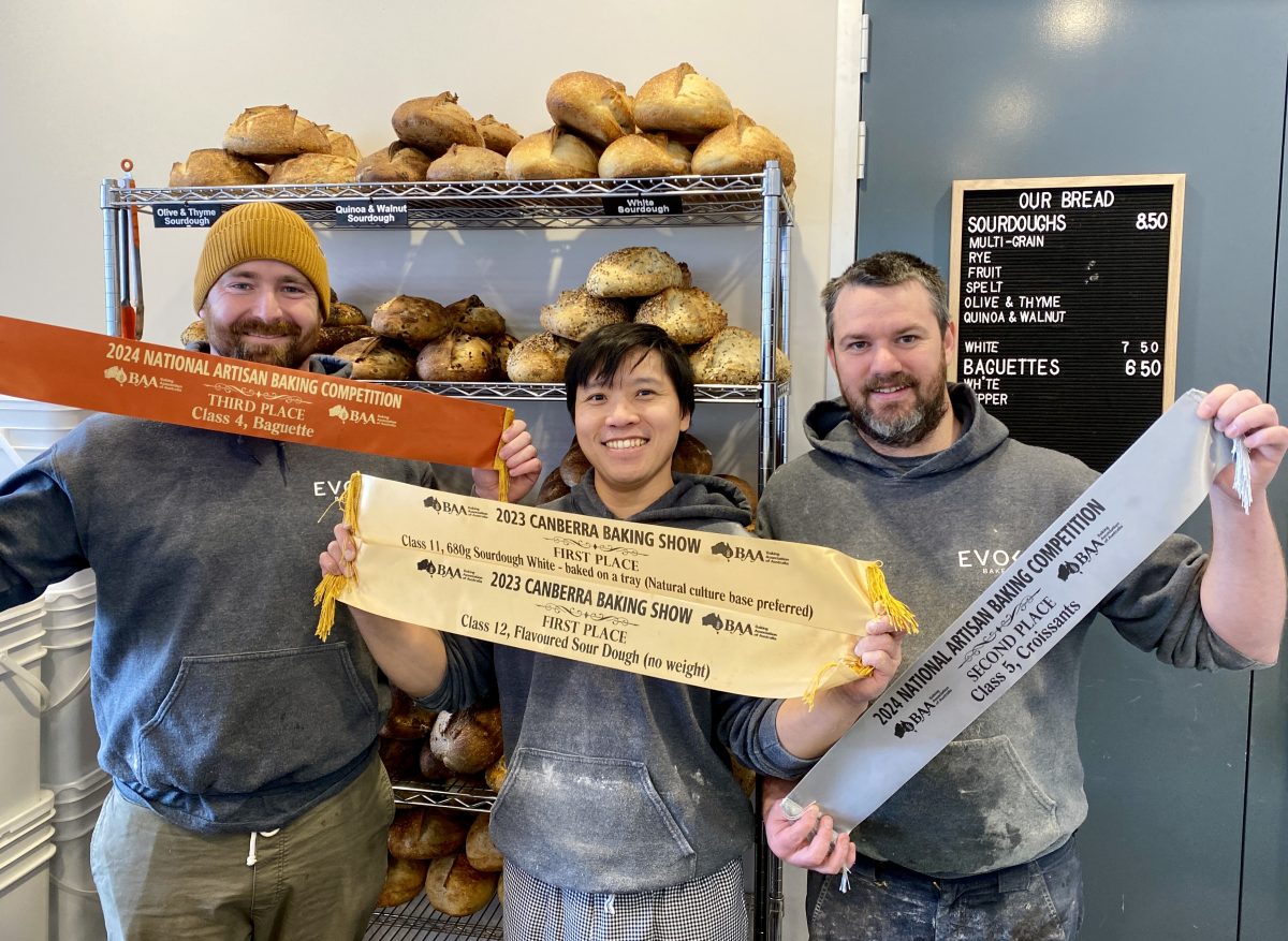 Three men dusted with flour hold gold, silver and bronze ribbons in front of bakery shelf.