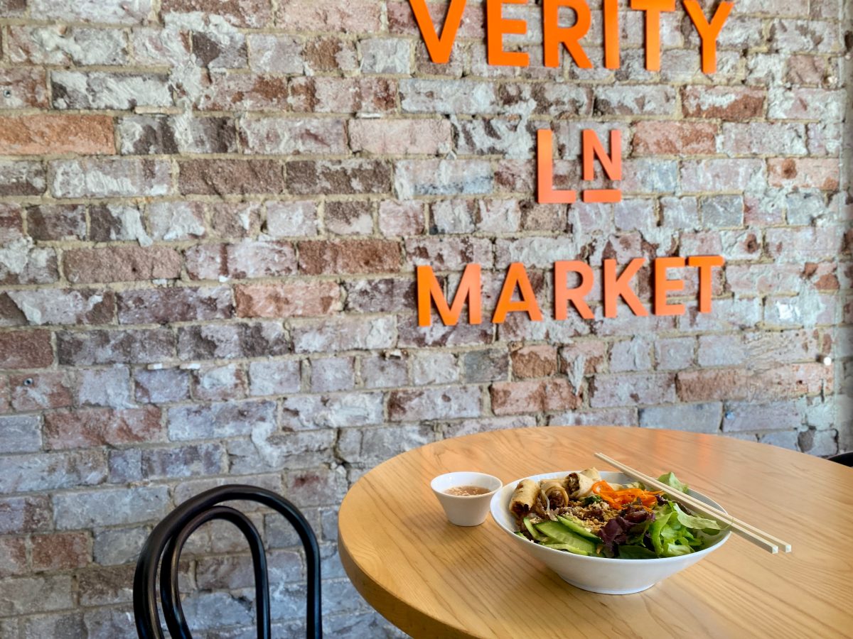 Bowl of salad and noodles in front of Verity Ln Market sign on brick wall.
