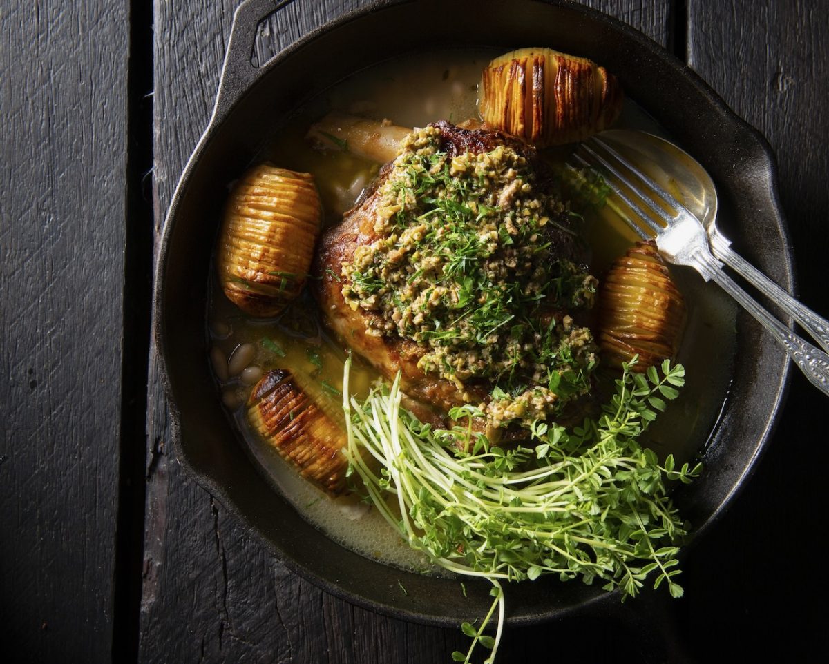 Dish of lamb shoulder in cast iron pan with hasselback potatoes and green garnishes. 