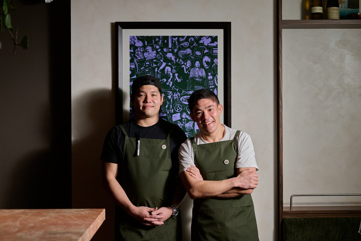 Mork and Ben wear green aprons and stand in front of purple and green artwork.