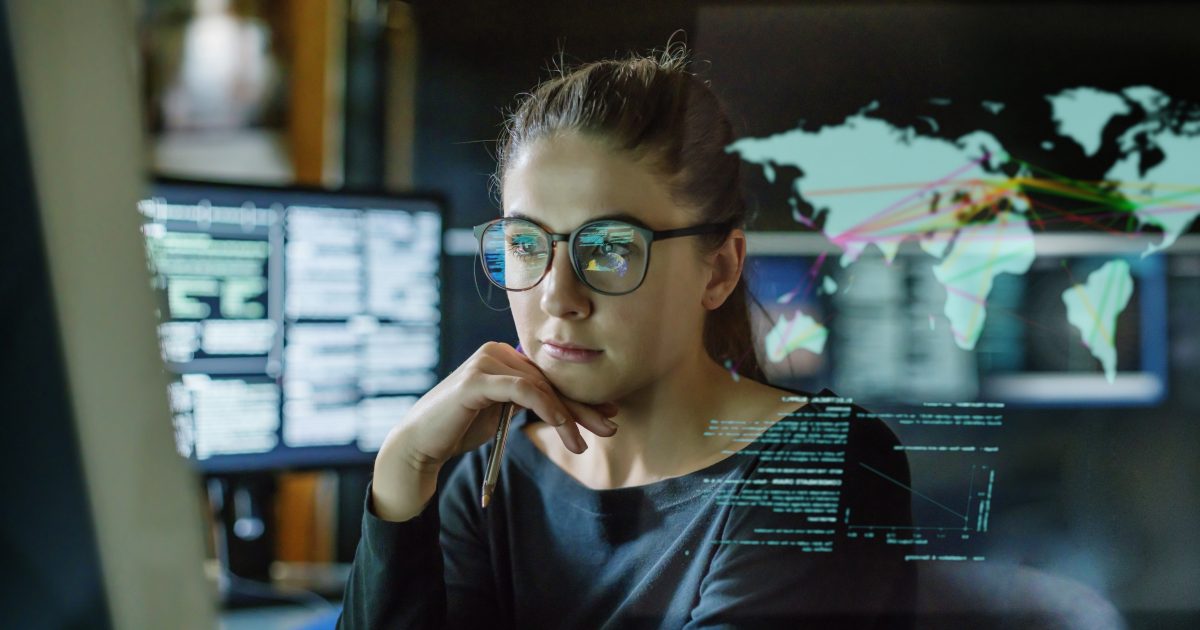 Stock image of a young woman, wearing glasses, surrounded by computer monitors in a dark office. In front of her there is a see-through displaying showing a map of the world with some data