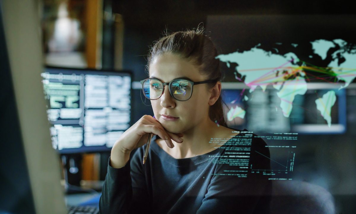 A young woman, wearing glasses, surrounded by computer monitors in a dark office. In front of her there is a see-through displaying showing a map of the world with other data