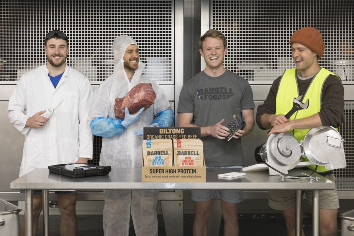 Four men stand at stainless steel work bench holding different tools and ingredients for making biltong like meat, a slicer, temperature probe and the finished product.