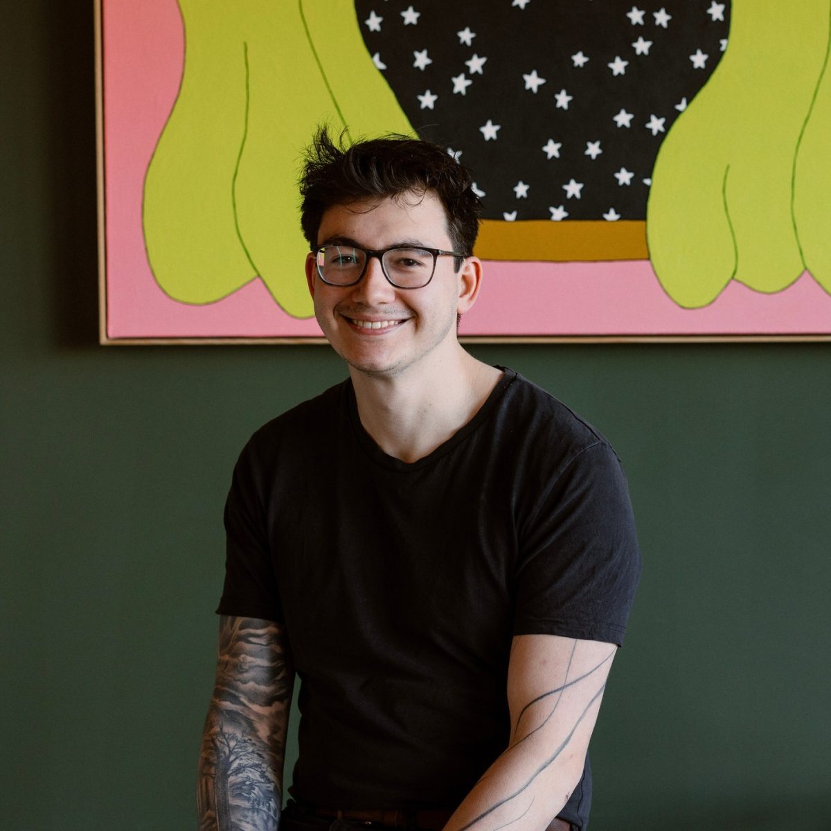 Reece wears glasses and a black t-shirt and sits in front of a quirky painting.