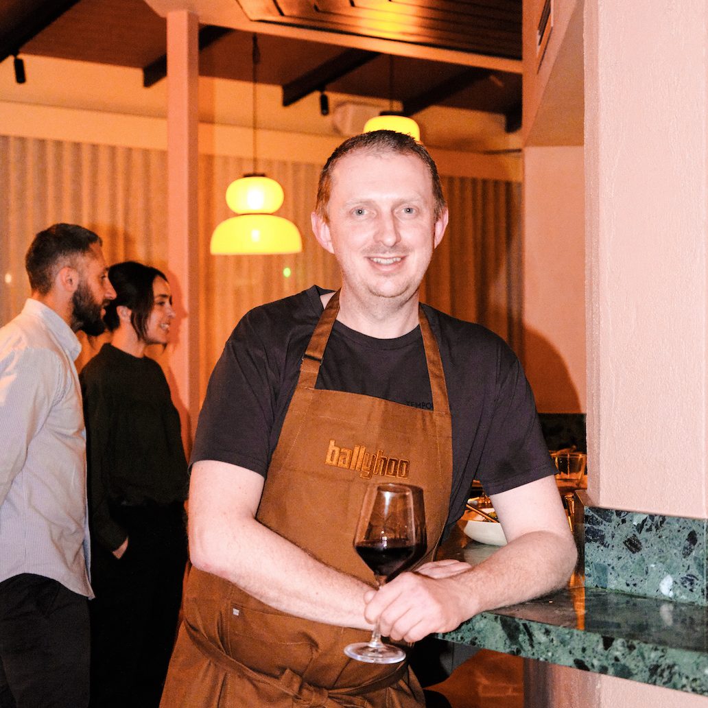 Dave wears a burnt-orange apron and holds a glass of red wine. 