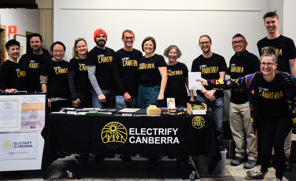 Group of 12 people wearing Electrify Canberra shirts standing around a table also brandishing the name.