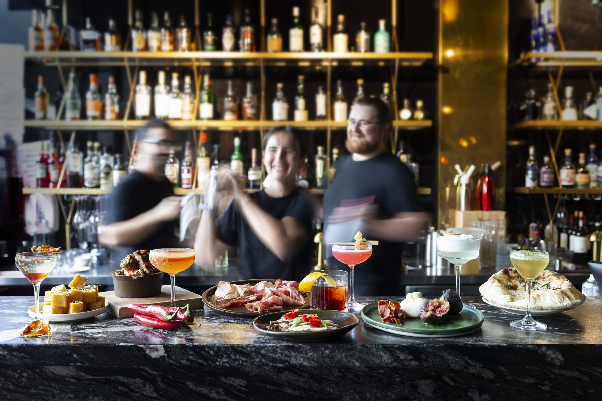 Many meals and drinks laid out on a bar in focus with movement blurred staff in background