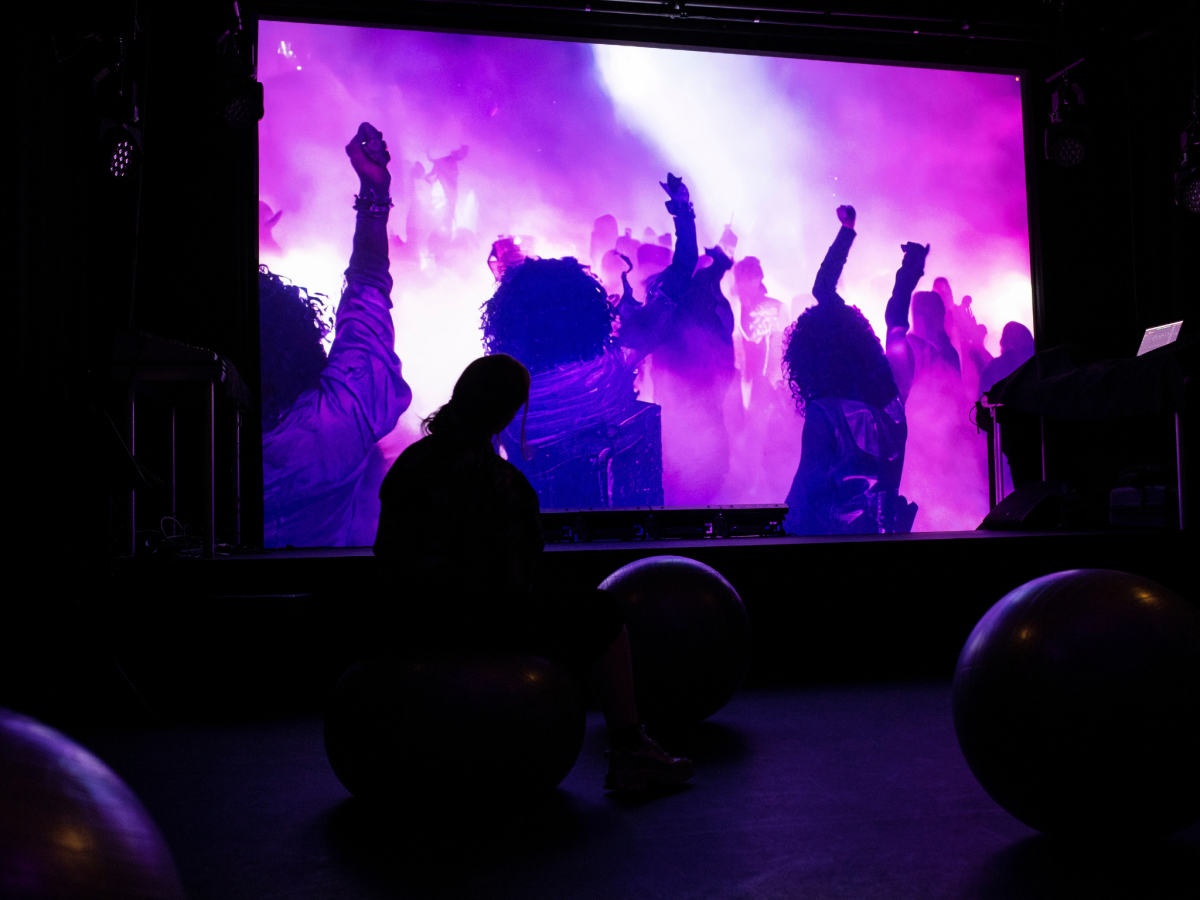 Silhouette of a person watching onscreen visuals of people dancing. The screen content is smoky and purple.