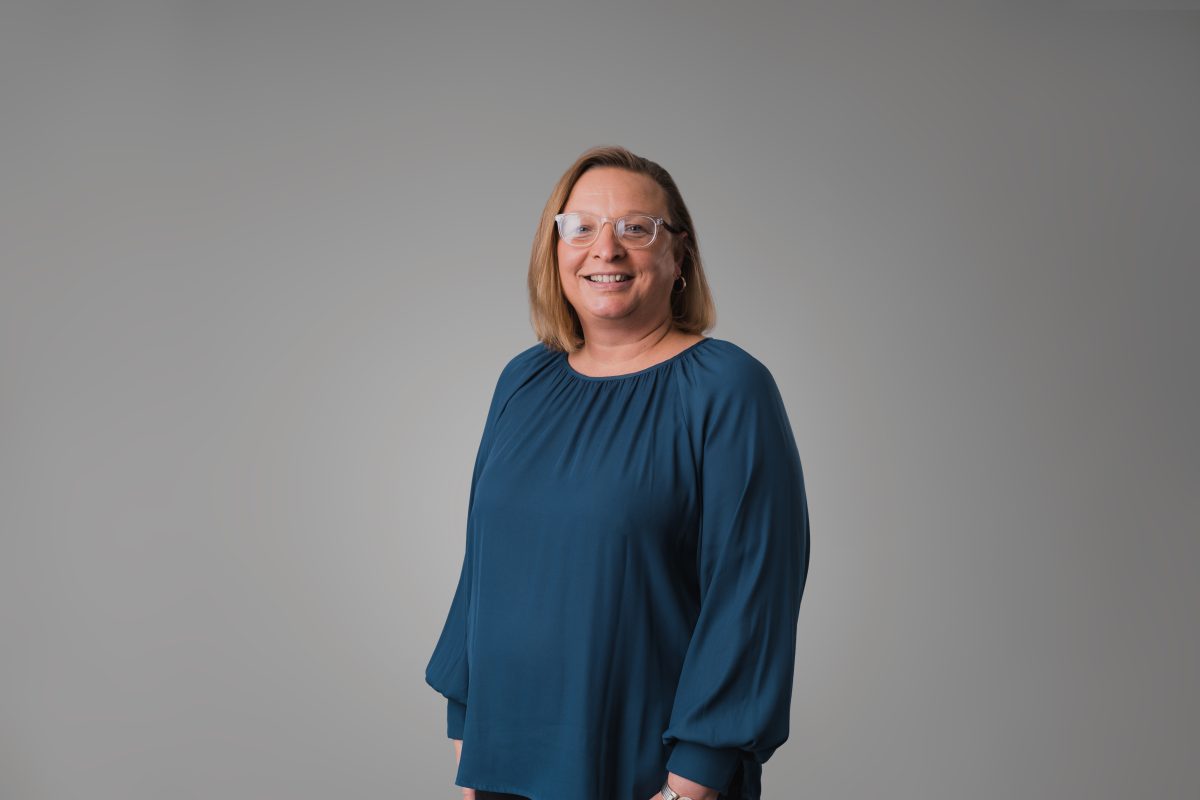 Woman standing in front of a grey background, She has blonde hair, is wearing glasses and a teal shirt.