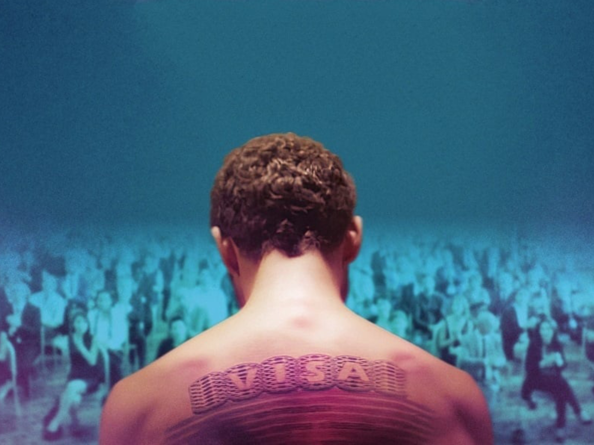 Promotional image from The Man Who Sold His Skin showing a man with a tattooed back sitting in front of a large audience. The tattoo says 'VISA'