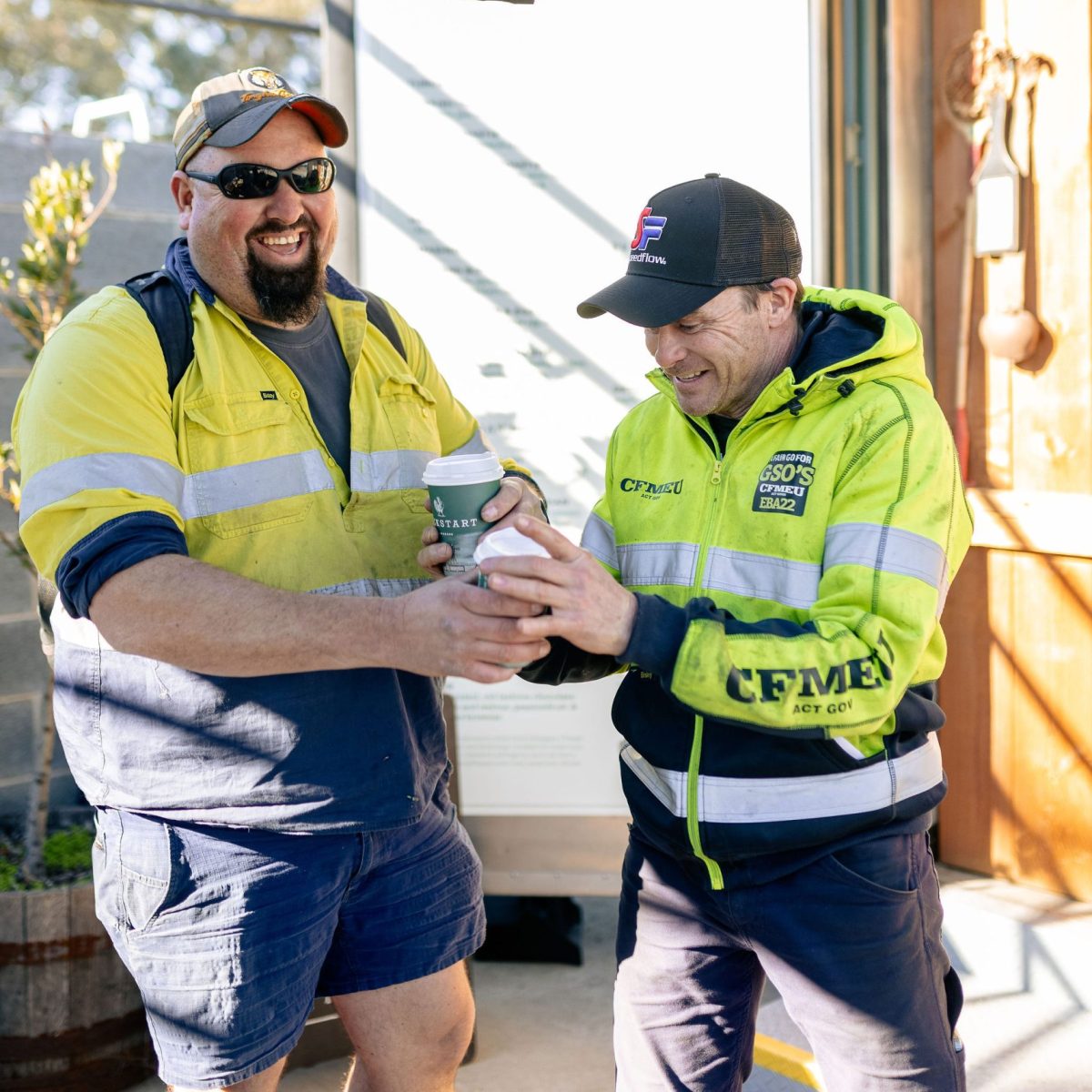 Two tradies hold coffee cups and smile.