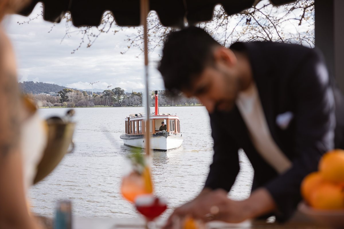 A bartender serves cocktails; in the background an old-fashioned boat sails on the lake.