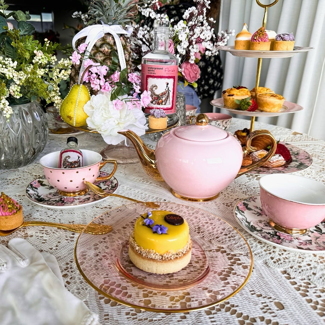 A table set for high tea with pink china, with a multi-layered yellow tart in the foreground.