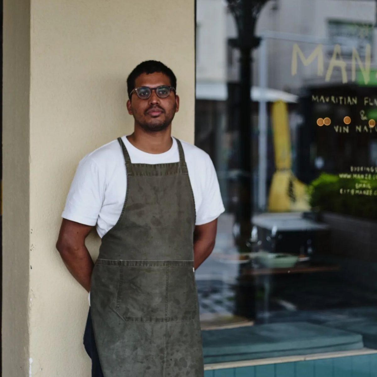 A chef in a white tshirt and apron leans against a wall next to a window which reads Manze.