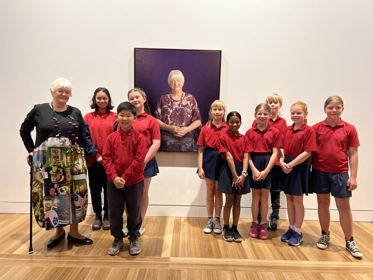 A group photo of school children in red uniforms in front of a portrait of Stephanie Alexander who stands next to the children.