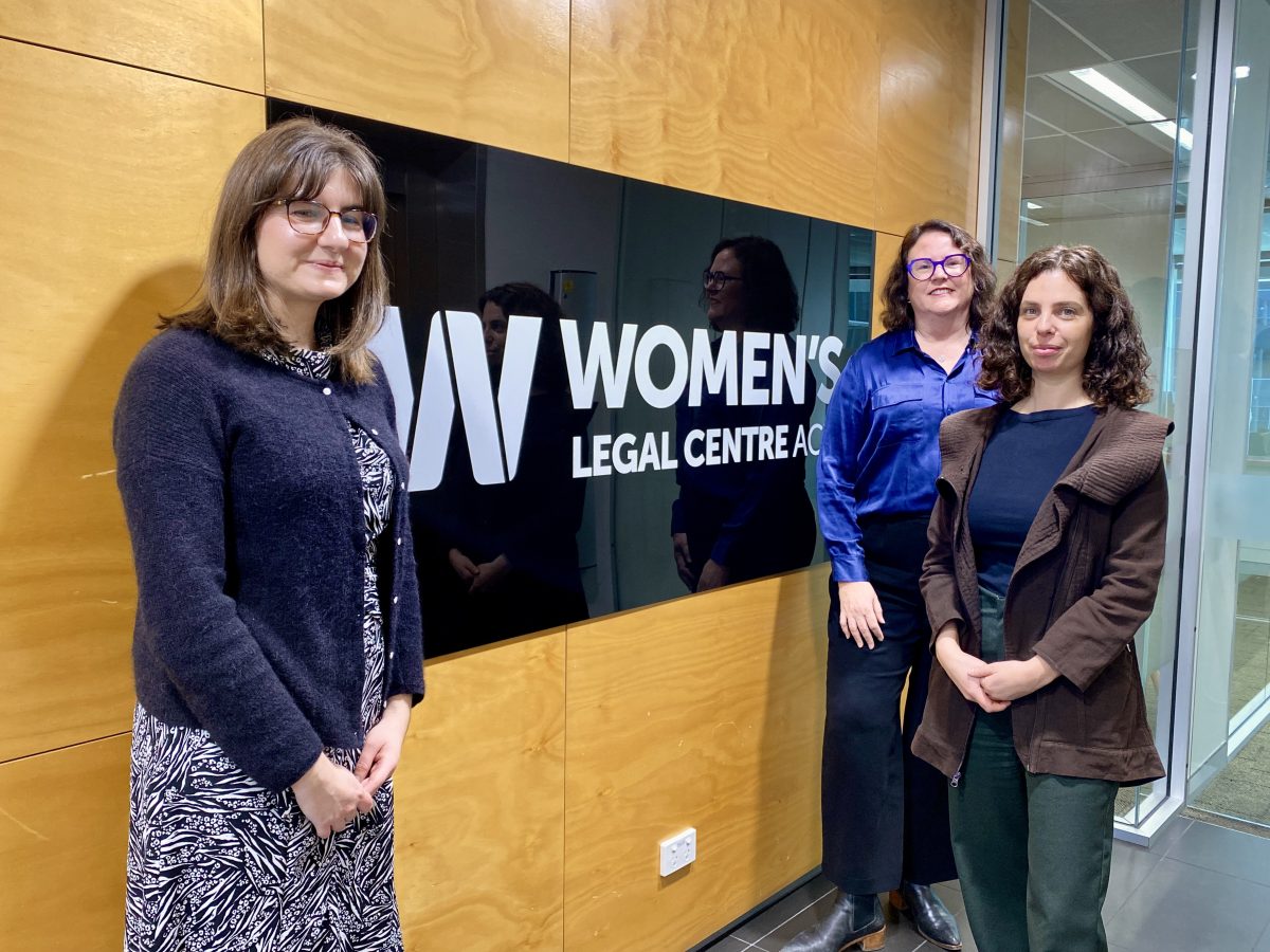 Three women stand beside a sign reading 'Women's Legal Centre ACT'