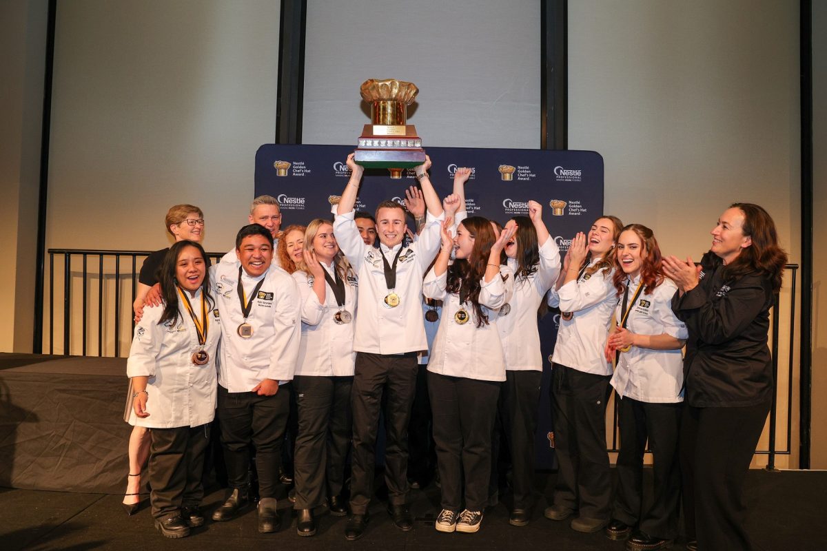 A group of young chefs in chef's uniforms pose as a group, the young man in the centre raises a trophy above his head.