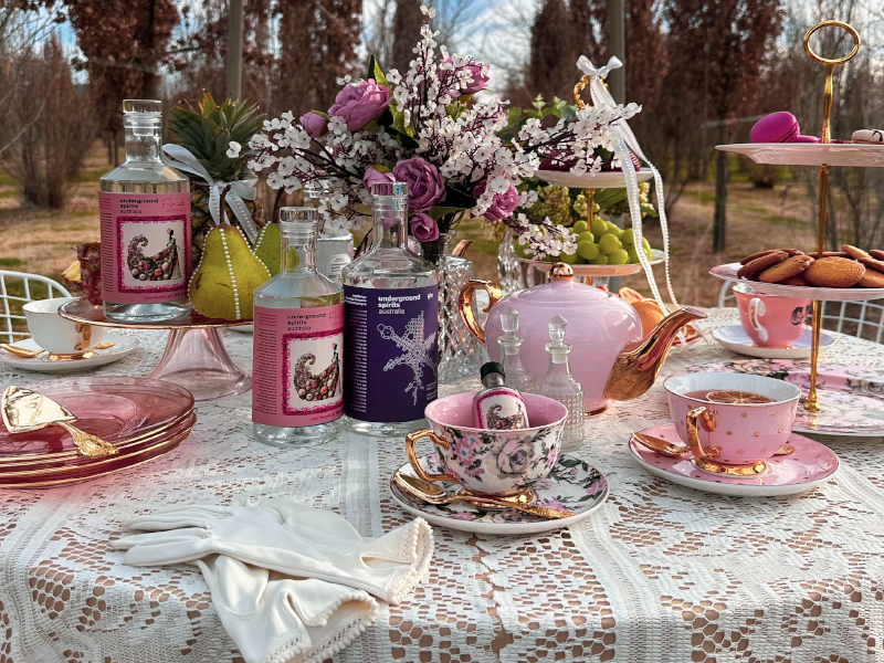 A table covered with lace tablecloth and set with pink and gold china, bottles of Underground Spirits gins, pastries and flowers. 