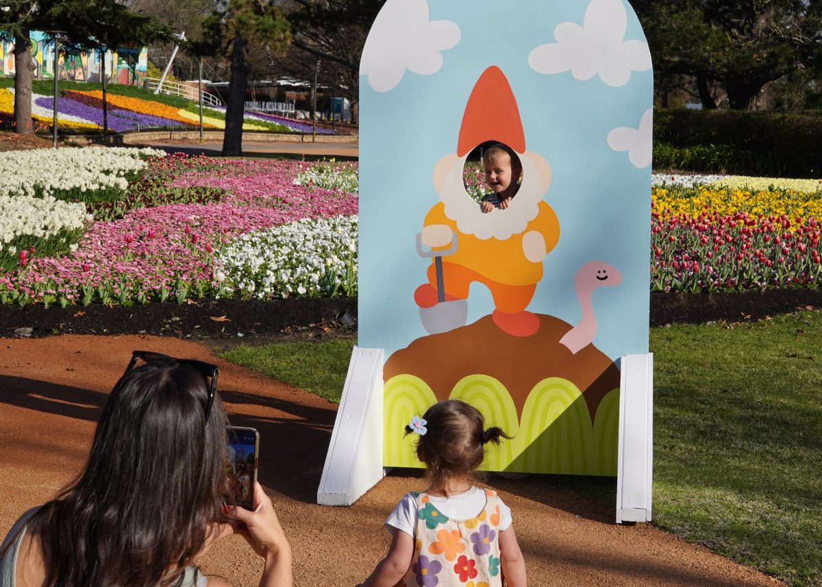 A person taking a photo of a kid posing with their head inside a hole painting of a gnome