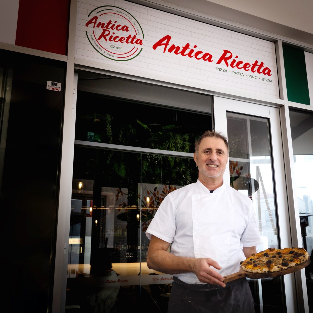 Chef holds a pizza outside restaurant with sign reading 'Antica Ricetta'