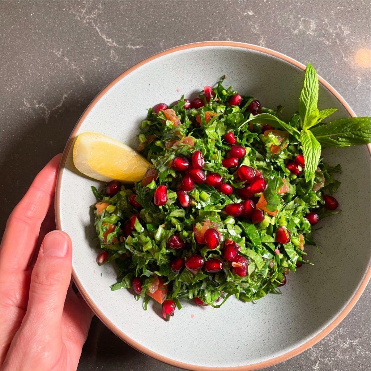 A hand holds a bowl of herby salad topped with pink pomegranate arils and a wedge of lemon.