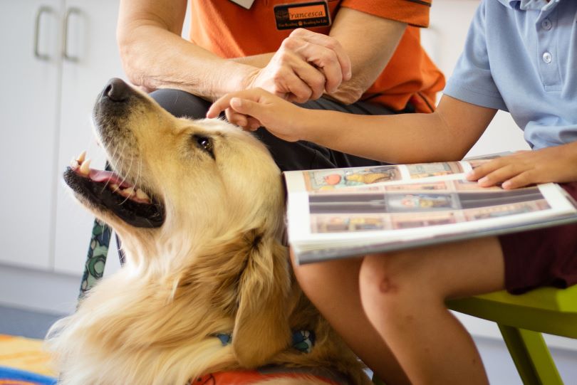 A photograph of a child reading with a dog and supervisor.