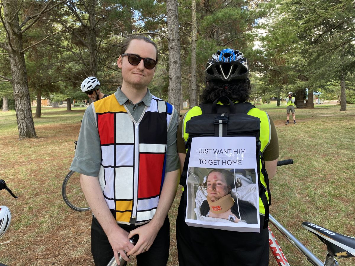 A man in a colourful vest stands next to someone with a picture of that man in a neck brace affixed to their back. The sign reads "I just want him to get home"