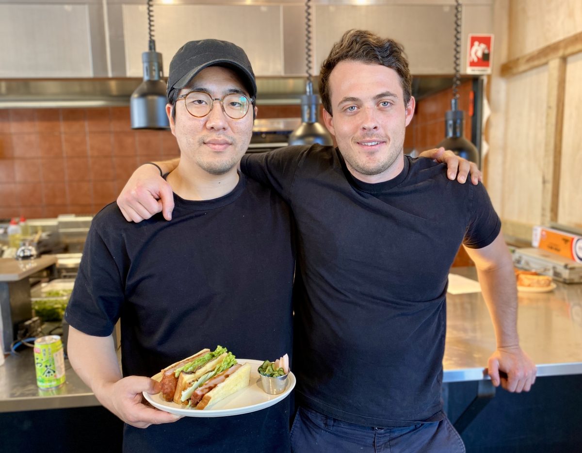 Two men in black stand in front of a kitchen holding a sandwich on a plate.