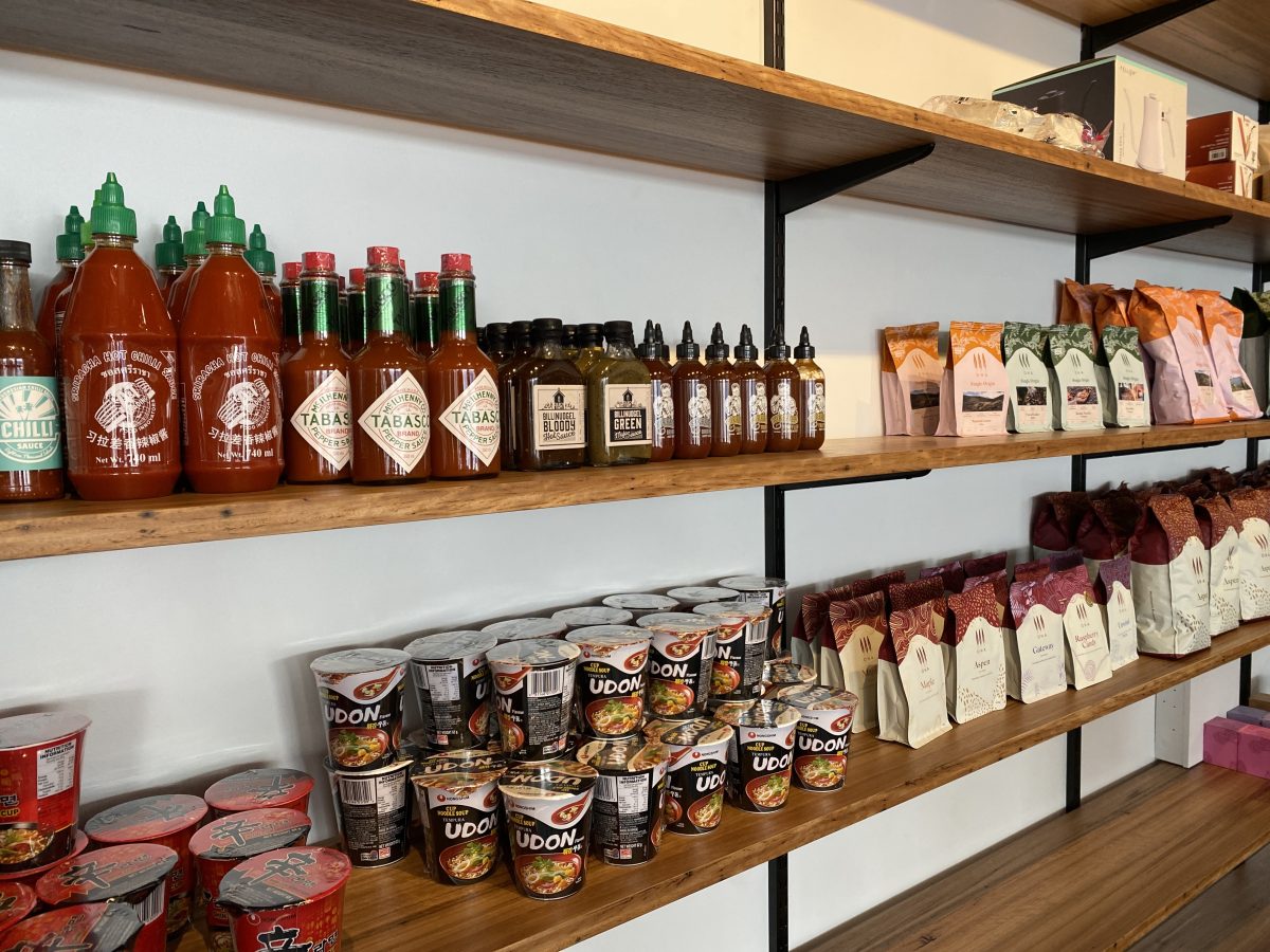 A retail shelf in a cafe displaying cup noodles, hot sauces and bags of ONA coffee.