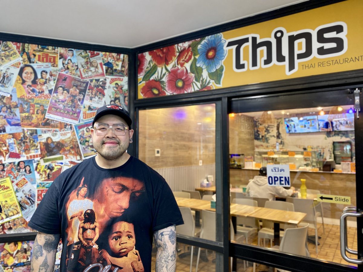 A man in a cap, glasses and shirt with images smiles outside Thip's Thai