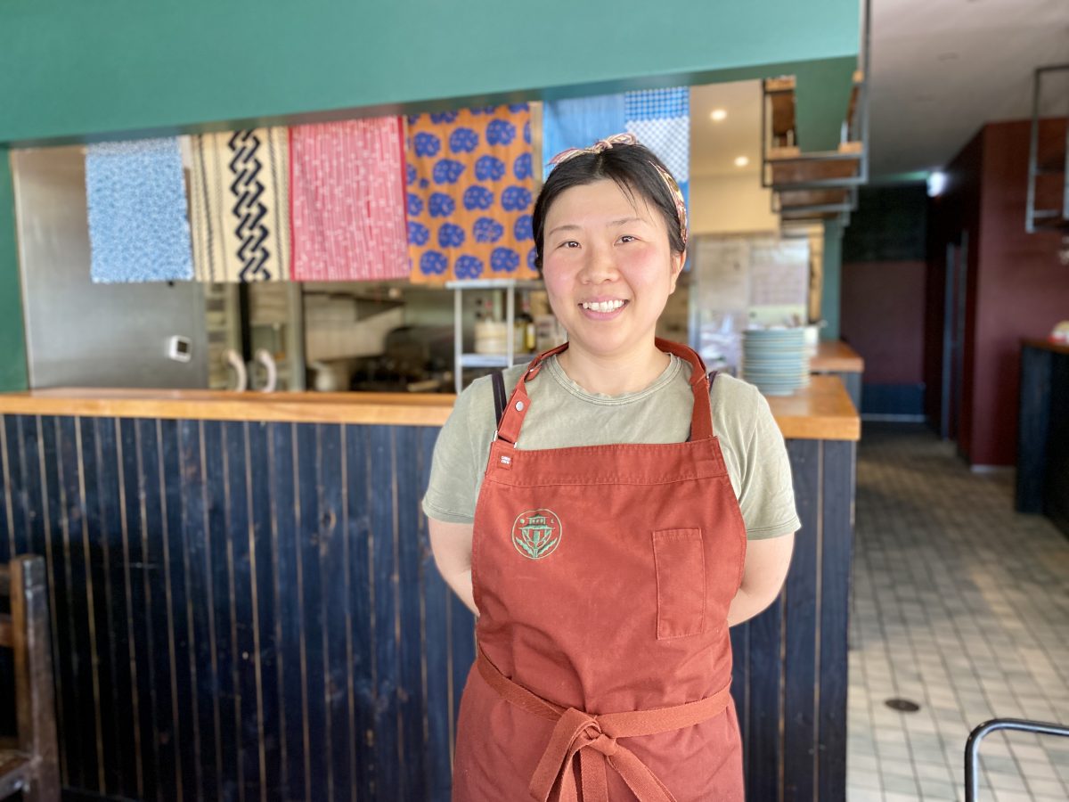 A woman with a headband wears an orange apron with green logo. 