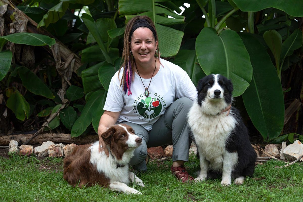 Woman with brown dreadlocks crouches in a garden with two dogs.