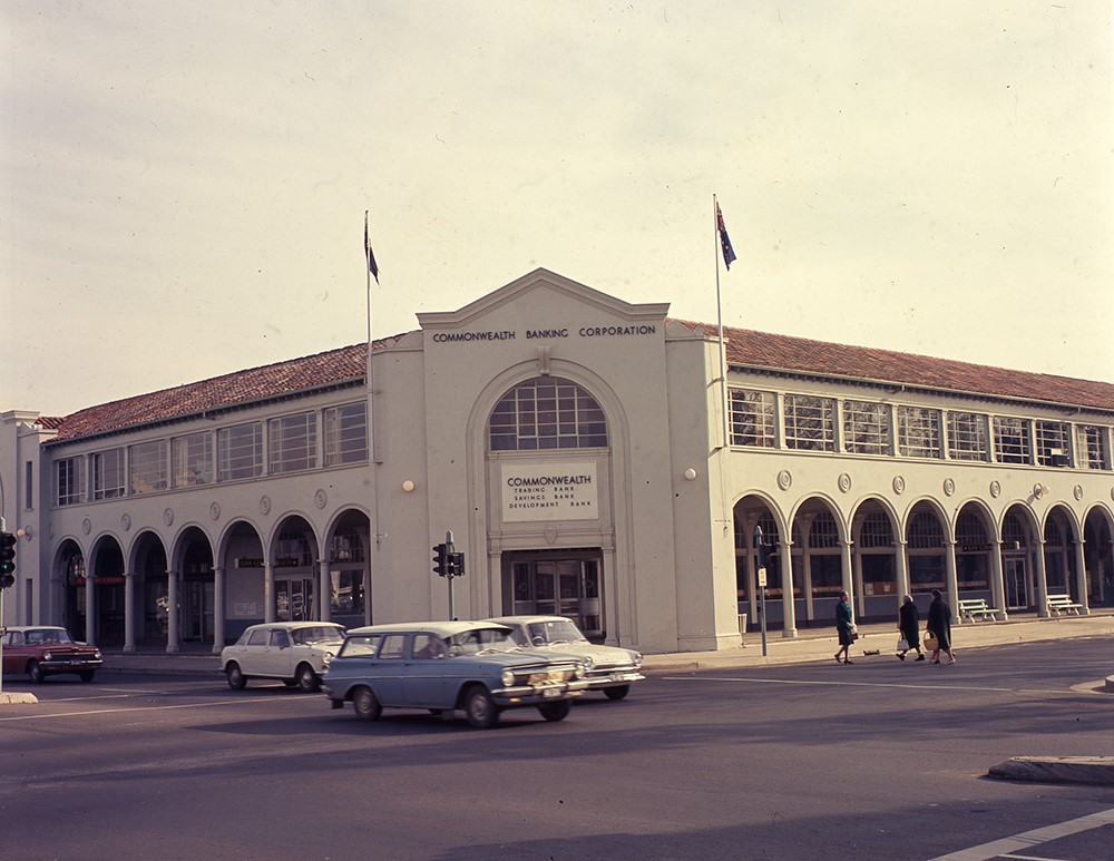 Black and white photo of corner building with cars on the road in front. 