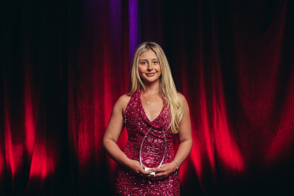 Ashleigh Bujaroski holding an award wearing a pink dress and standing in front of a red curtain