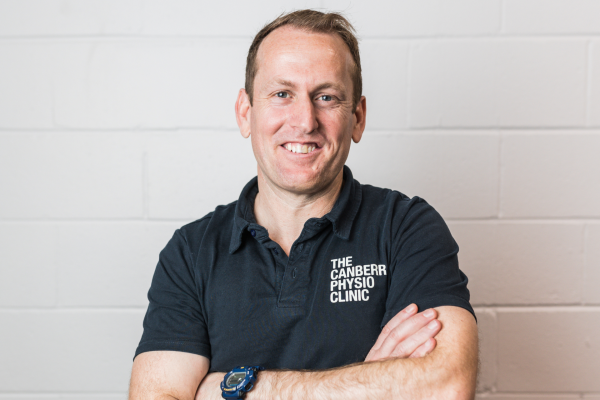 Toby Conroy wearing Canberra Physio Clinic uniform with arms crossed standing in front of a white brick wall