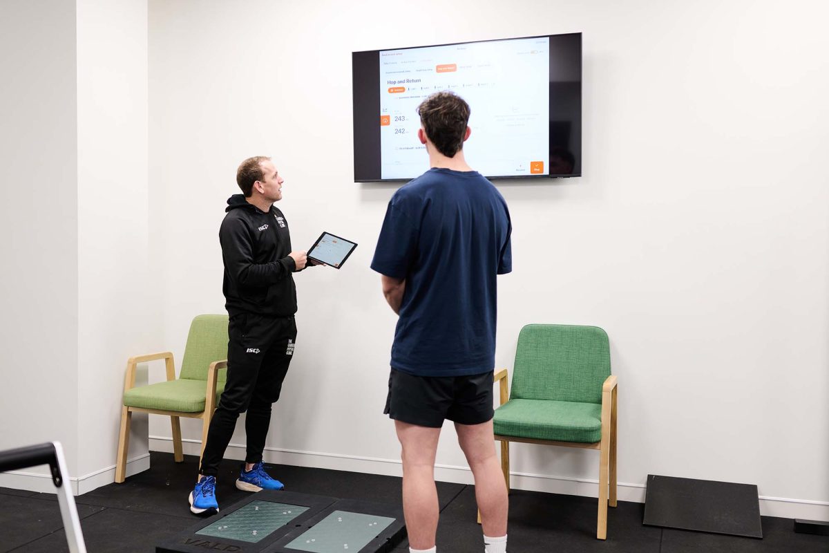 Toby Conroy and another man stand n a room next to VALD Equipment looking at a TV