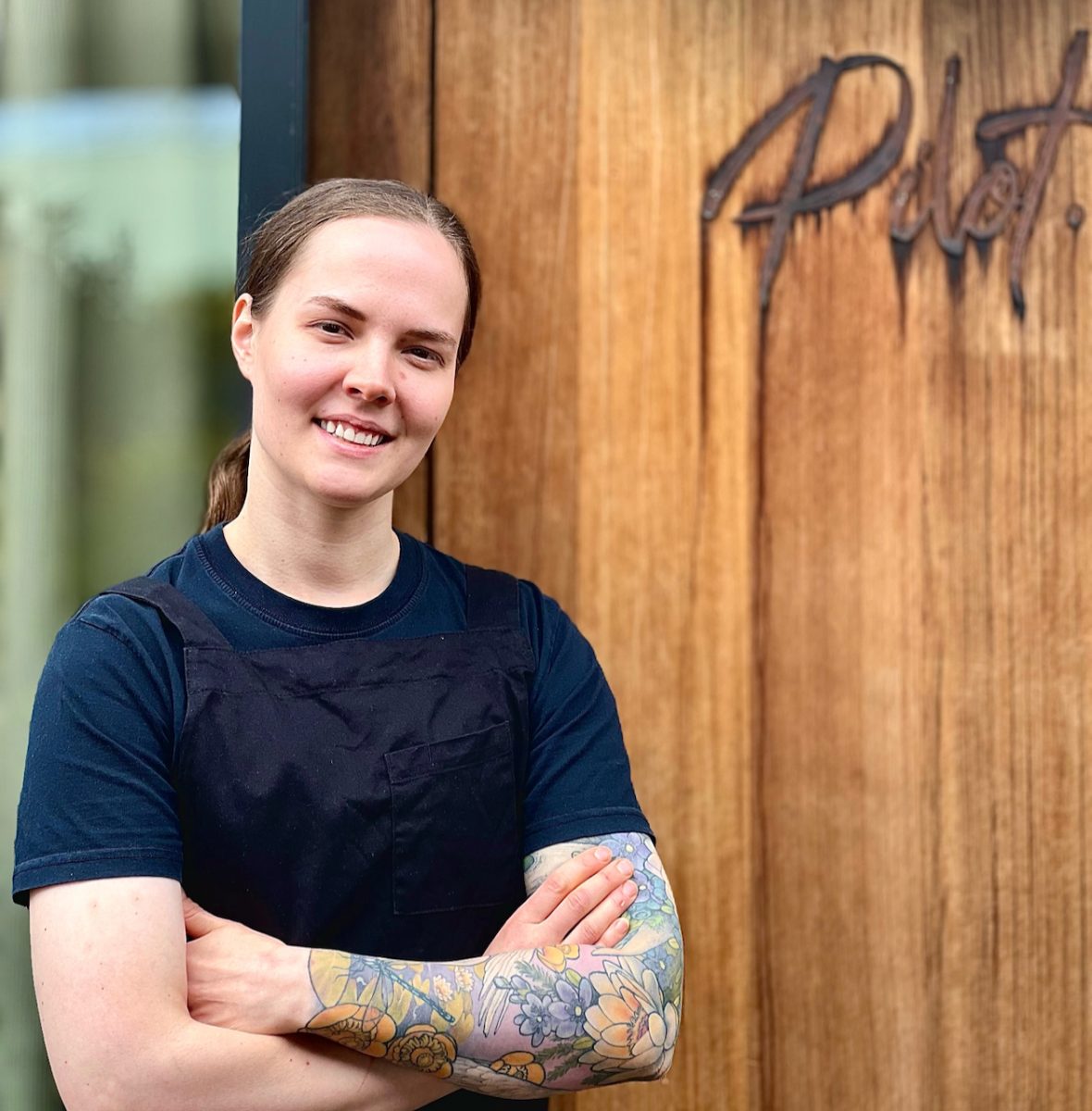 A young woman with a sleeve of tattoos smiles with crossed arms in front of a wooden door which reads 'Pilot'.