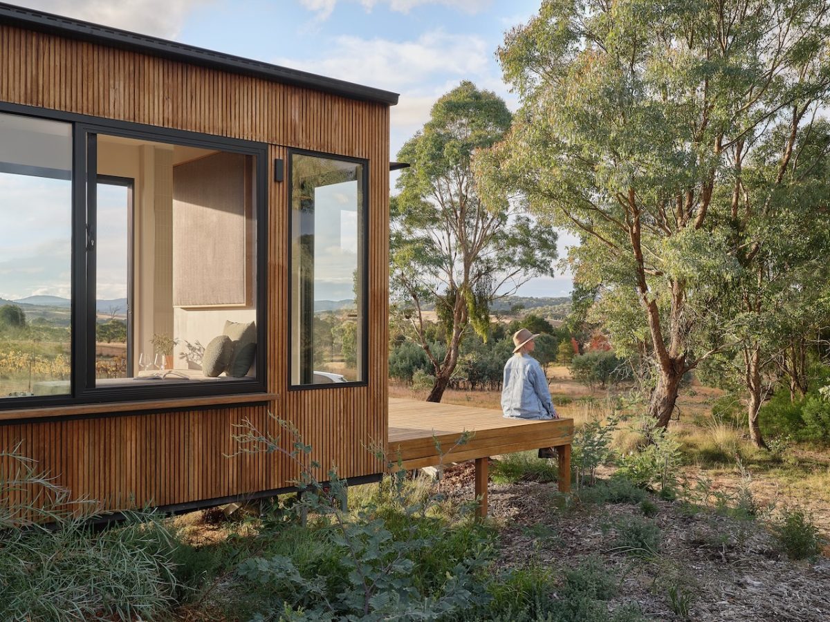 Woman sits on the deck of a wooden tiny house in the bush. 