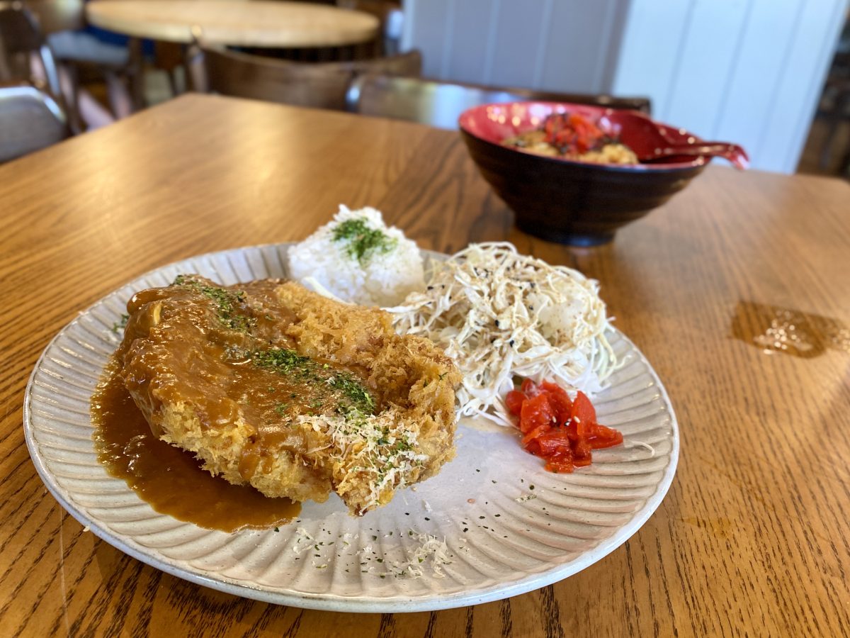 A plate with a crumbed and fried piece of pork covered in a gravy, served alongside red pickles, shredded cabbage and rice. 