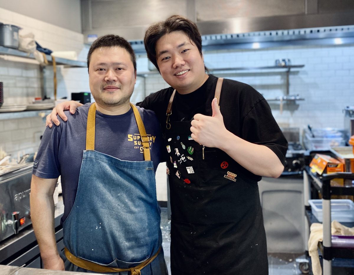 Two men in aprons stand in a kitchen, one gives a thumbs up sign.