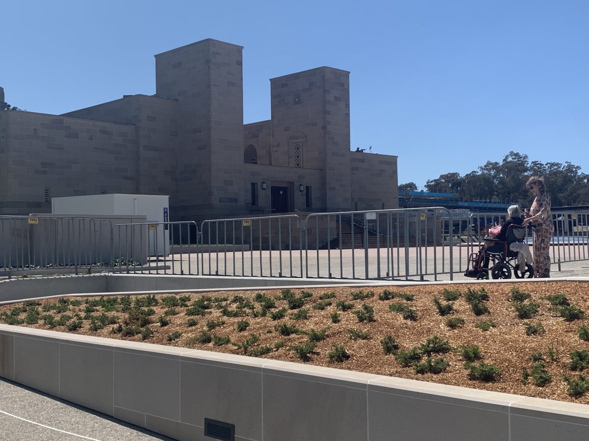 An elderly woman in a wheelchair with her carer behind her sitting before the Australian War Memorial with two snipers on each side of the roof.