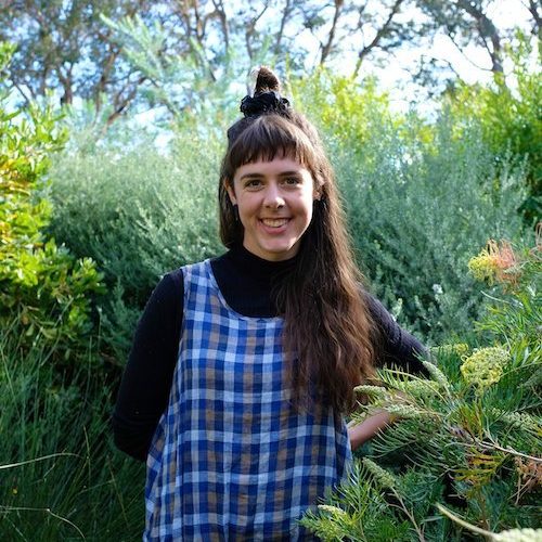 A women with fringe, high bun and plaid smock stands in a garden.