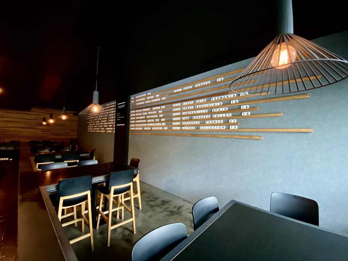 A minimalist restaurant interior with black bar tops, stools, a menu on the wall and warm exposed lighting