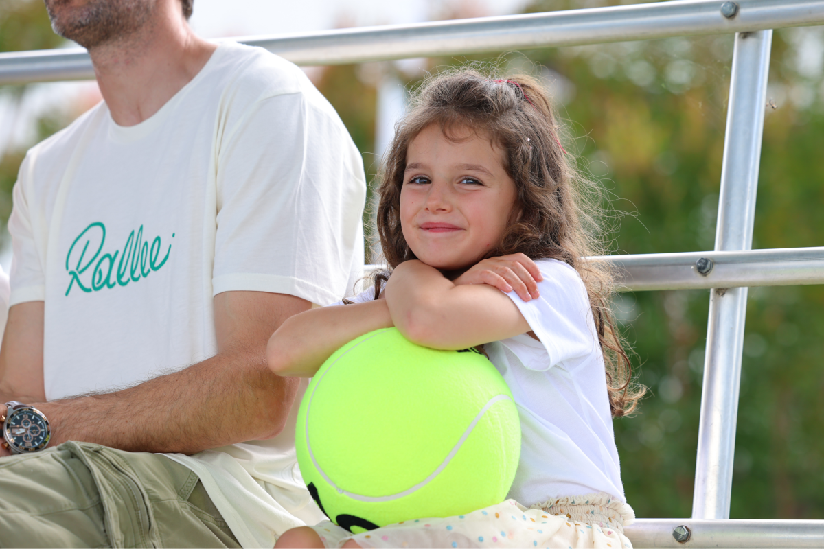 a young child holds a large tennis ball sitting in a grandstand next to an adult