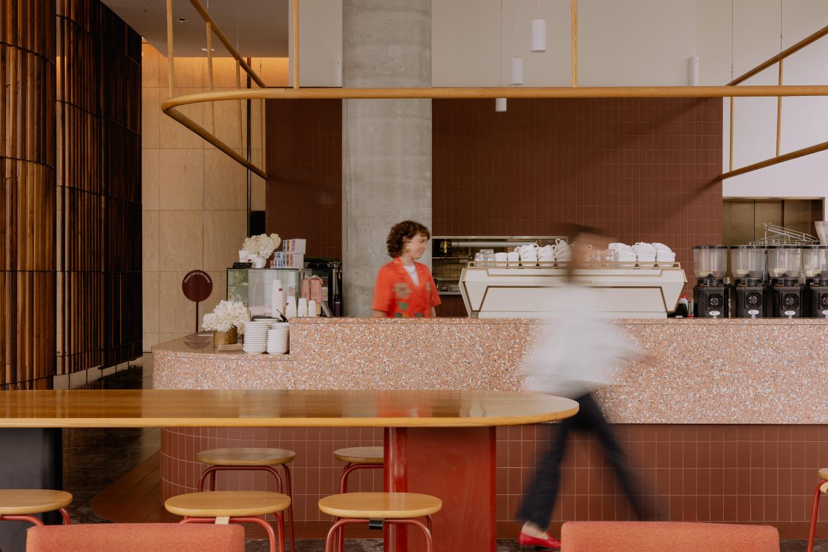 A person in white, blurred with speed, walks past a barista in a red-themed cafe.