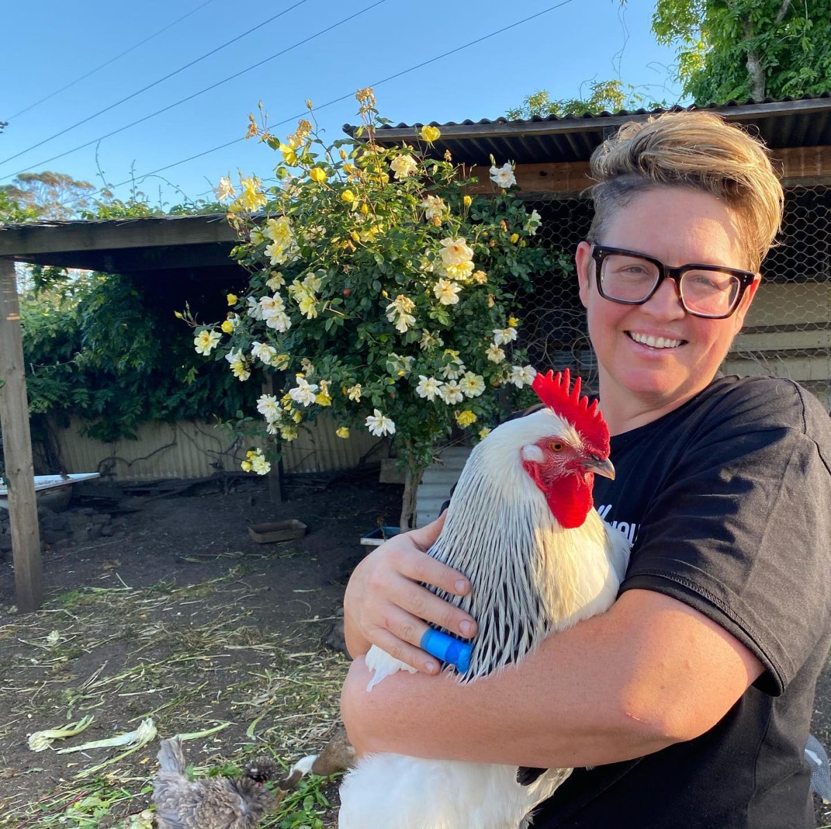 A woman with large glasses holds a rooster, there is a flowering plant behind her.