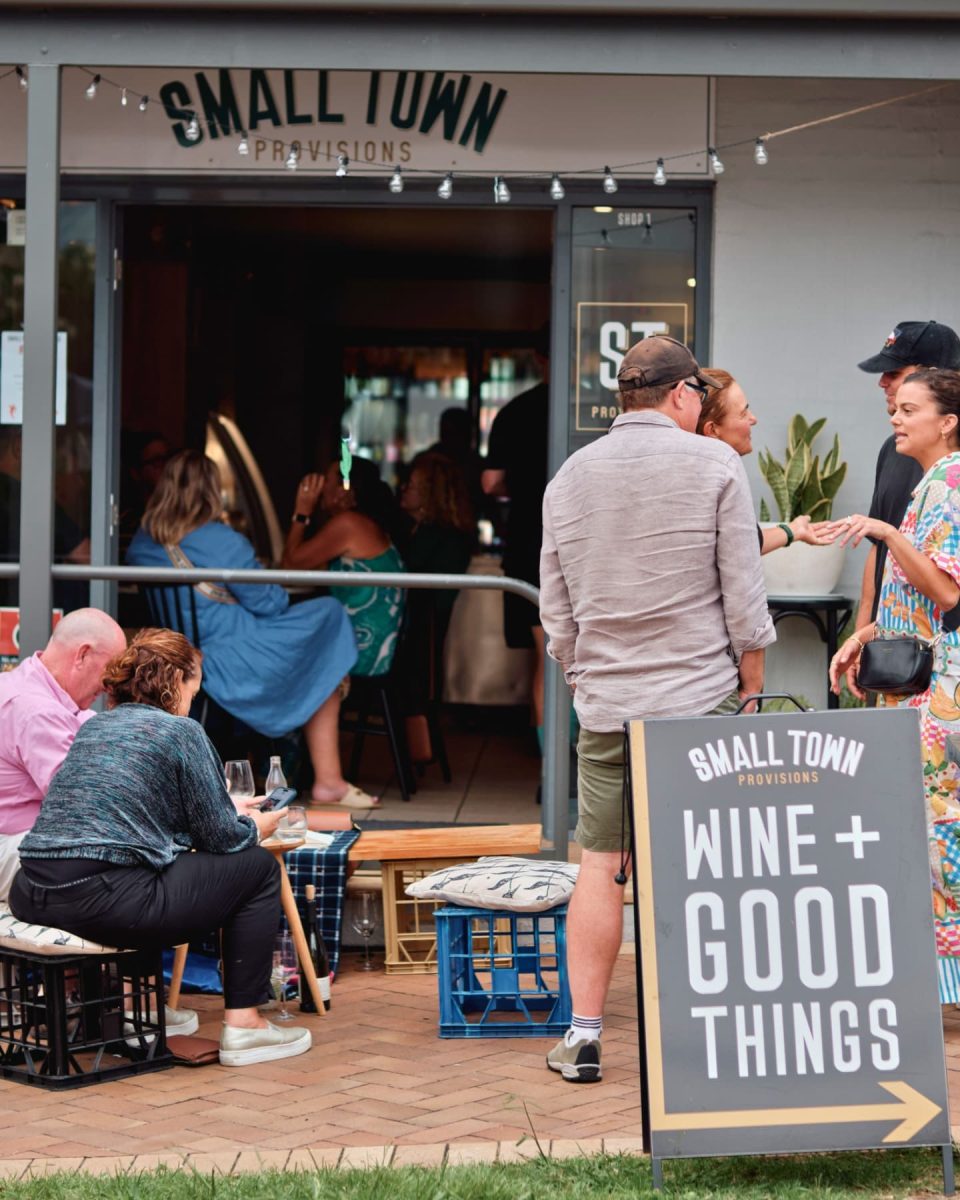 An A-frame sign reading 'Wine + Good Things' outside a store filled with people drinking.