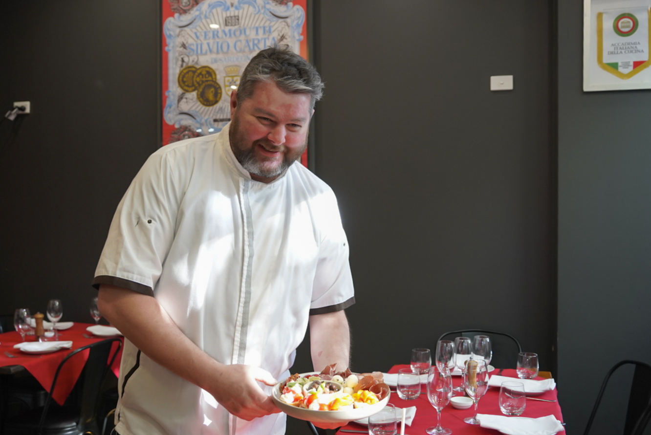 A chef stands in a restaurant holding a plate of food.